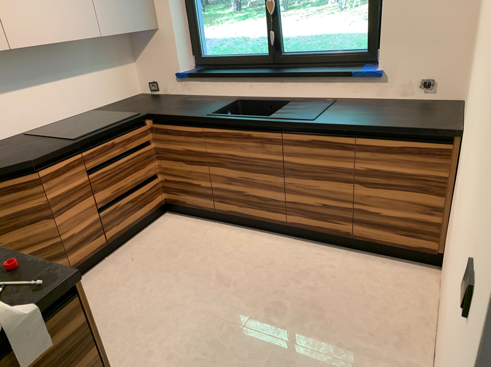 Nowoczesna kuchnia z frontami szafek w kolorze ciemnego drewna ze wzorem zebry, czarnym blatem i zlewem, białe ściany i podłoga z połyskiem, okno z widokiem na zieleń.