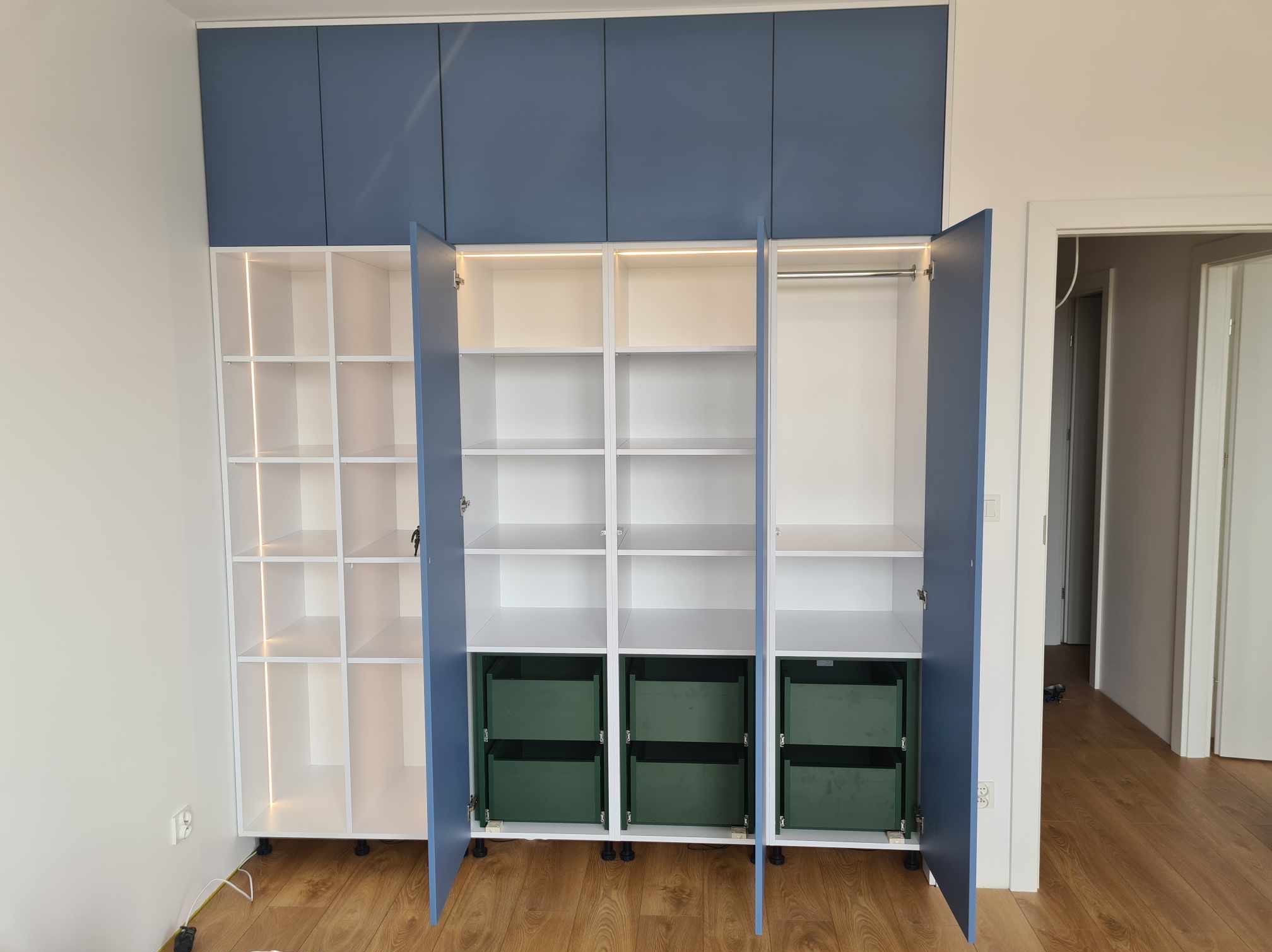 Szafa wnękowa z niebieskimi drzwiami, białymi półkami i zielonymi szufladami, podświetlana od wewnątrz