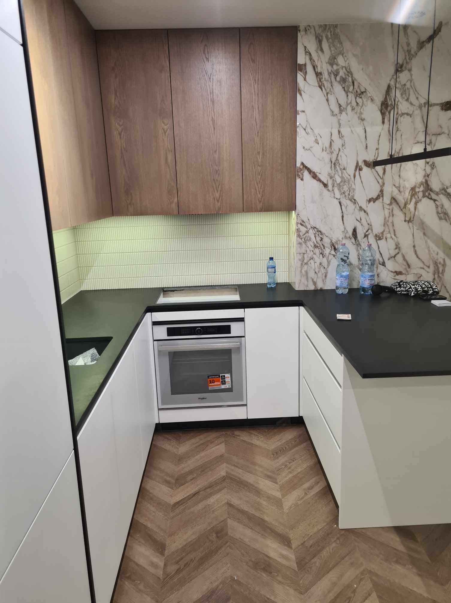 Nowoczesna kuchnia z drewnianymi szafkami górnymi, białymi szafkami dolnymi i blatem z czarnego kamienia, marmurowa ściana w tle, parkiet w jodełkę na podłodze.