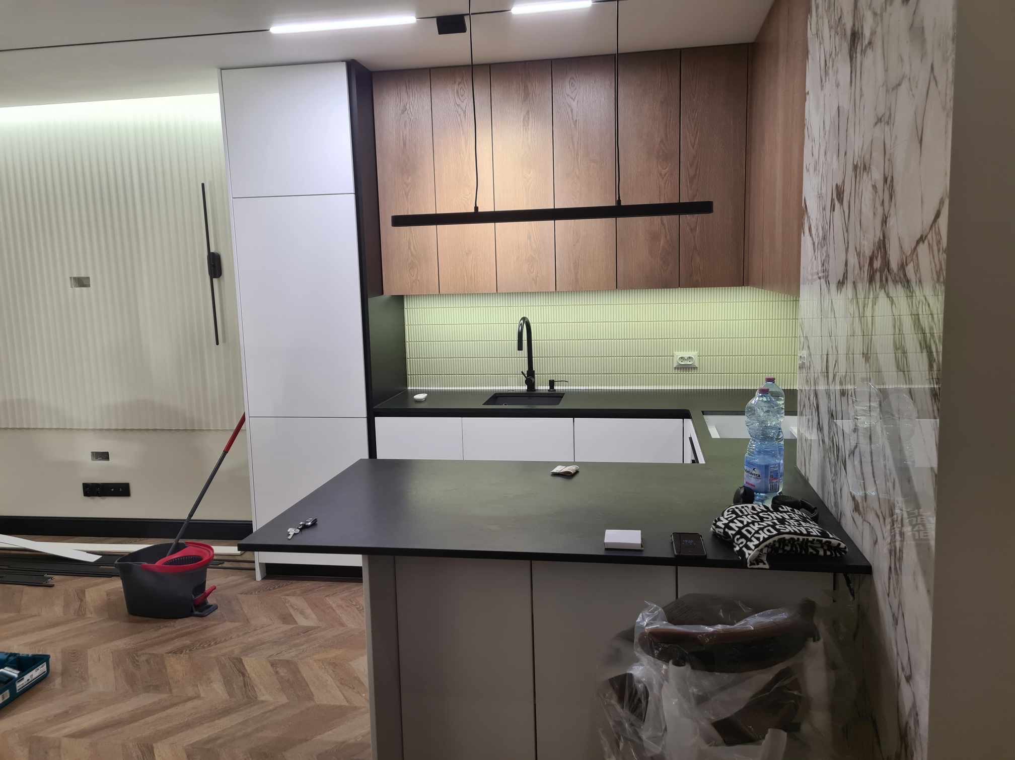 Montaż nowoczesnej kuchni: widoczne szafki wiszące z drewnianymi frontami, białe szafki stojące, czarny blat, oświetlenie liniowe i fragment ściany z marmurowym wzorem. Na pierwszym planie wyspa...