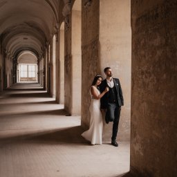 Mariusz Kędzierski - Para młoda: panna młoda w białej sukni i pan młody w czarnym garniturze, pozują w długim korytarzu z łukami i kolumnami, oświetlonym naturalnym światłem wpadającym z okien na końcu korytarza.