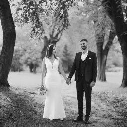 Mariusz Kędzierski - Czarno-białe zdjęcie pary młodej trzymającej się za ręce na tle alei drzew. Panna młoda w prostej sukni z bukietem, pan młody w garniturze z poszetką. Romantyczna sceneria.