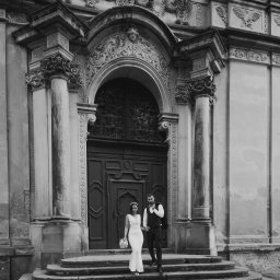 Mariusz Kędzierski - Czarno-białe zdjęcie pary młodej schodzącej po kamiennych schodach zabytkowego budynku z bogato zdobionym portalem.