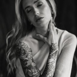 Mariusz Kędzierski - Czarno-białe, artystyczne ujęcie kobiety z tatuażami na rękach, z zamkniętymi oczami, w swetrze, delikatnie dotykającej szyi. Subtelne światło i głębia ostrości.