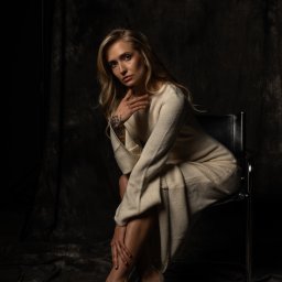 Mariusz Kędzierski - Portret kobiety w kremowej sukience, siedzącej na krześle na ciemnym tle. Widoczny tatuaż na ręce i beżowe szpilki. Subtelne oświetlenie podkreśla rysy twarzy.