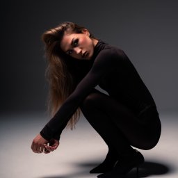 Mariusz Kędzierski - Kobieta w czarnym kombinezonie pozuje w kucki na szarym tle, oświetlona punktowym światłem, z uwagą na jej wyrazistym makijażu i luźnych włosach.