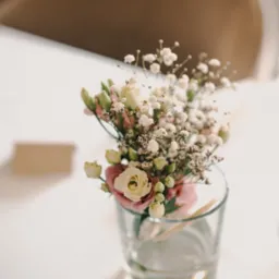 Delikatny bukiecik z białych i różowych kwiatów w szklanym wazonie na stole z kartą w tle, prawdopodobnie dekoracja stołu weselnego w ciepłym oświetleniu.