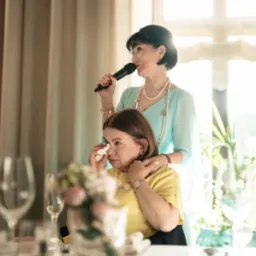 Kobieta z mikrofonem przemawia, a druga kobieta ociera łzy wzruszenia. Ujęcie z bliska, w tle okno i zasłony, widoczne kieliszki i bukiet kwiat&oacute;w na stole.