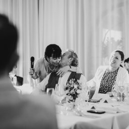 Mariusz Kędzierski - Czarno-białe zdjęcie z wesela: kobieta z mikrofonem całuje pana młodego przy stole, goście weselni obserwują z uśmiechem. Romantyczna atmosfera.