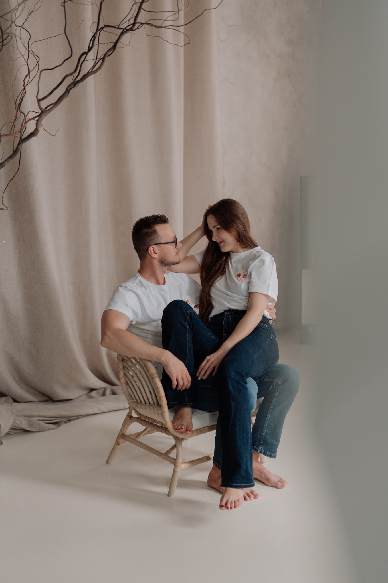 Para ubrana w białe koszulki i dżinsy, siedząca na wiklinowym krześle, kobieta na kolanach mężczyzny, oboje boso, patrzą na siebie z uśmiechem, neutralne tło z beżową zasłoną i gałęzią.