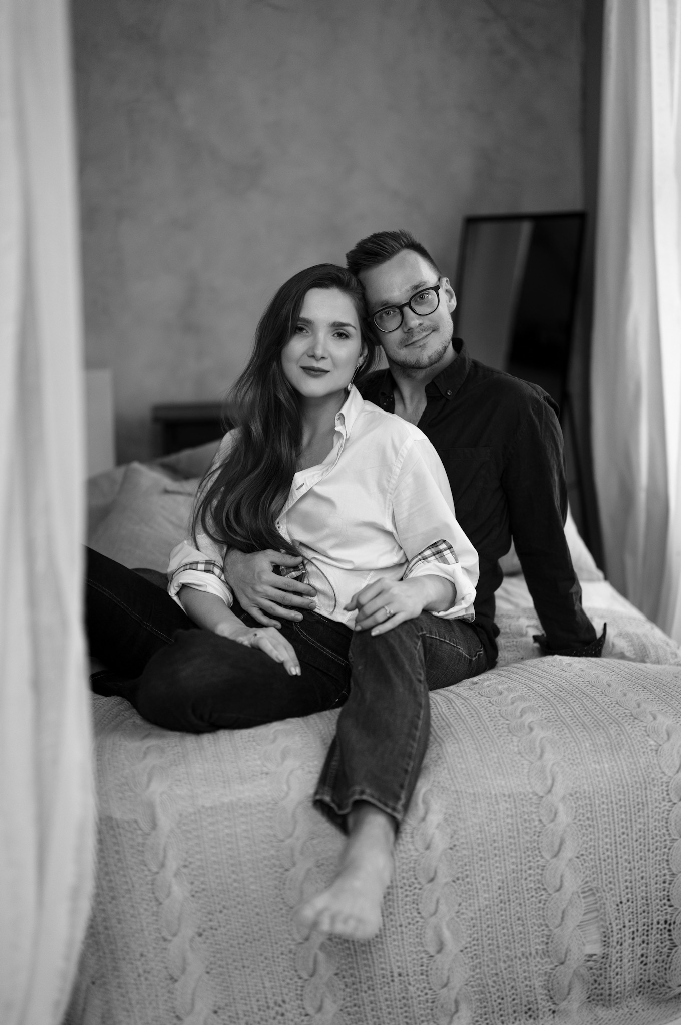 Czarno-białe zdjęcie narzeczonej i narzeczonego siedzących na łóżku z dzianinową narzutą, mężczyzna w okularach obejmuje kobietę w białej koszuli.