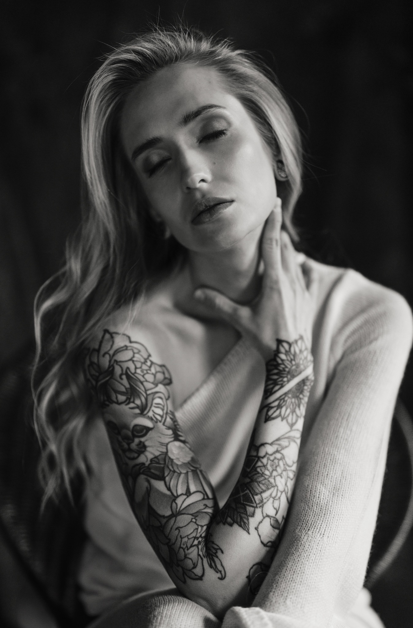 Czarno-białe, artystyczne ujęcie kobiety z tatuażami na rękach, z zamkniętymi oczami, w swetrze, delikatnie dotykającej szyi. Subtelne światło i głębia ostrości.