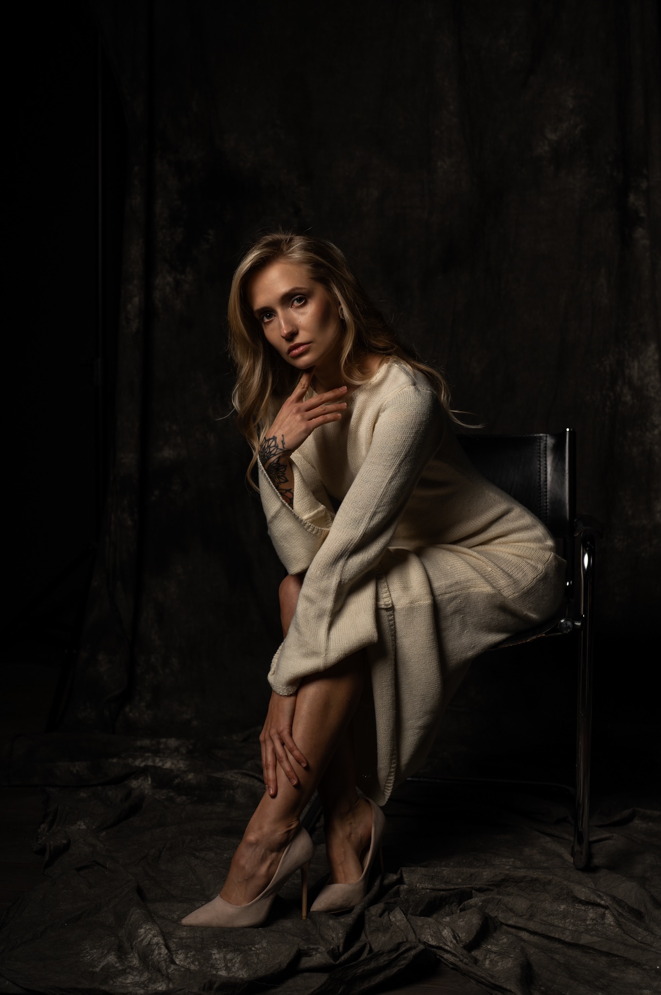 Portret kobiety w kremowej sukience, siedzącej na krześle na ciemnym tle. Widoczny tatuaż na ręce i beżowe szpilki. Subtelne oświetlenie podkreśla rysy twarzy.