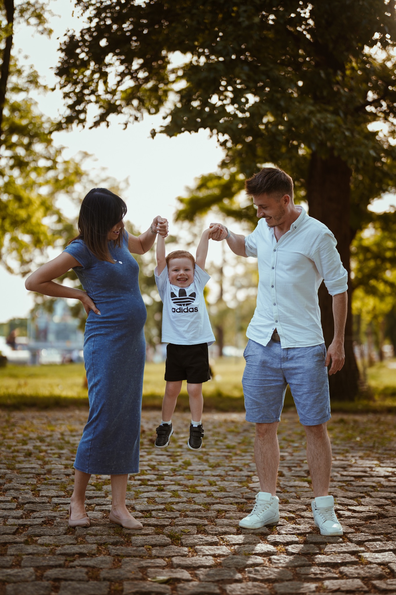Sesja rodzinna we Wrocławiu: Uśmiechnięty chłopiec podnoszony przez rodziców na tle parku. Mama w ciąży, tata w białej koszuli. Ciepłe, naturalne światło.