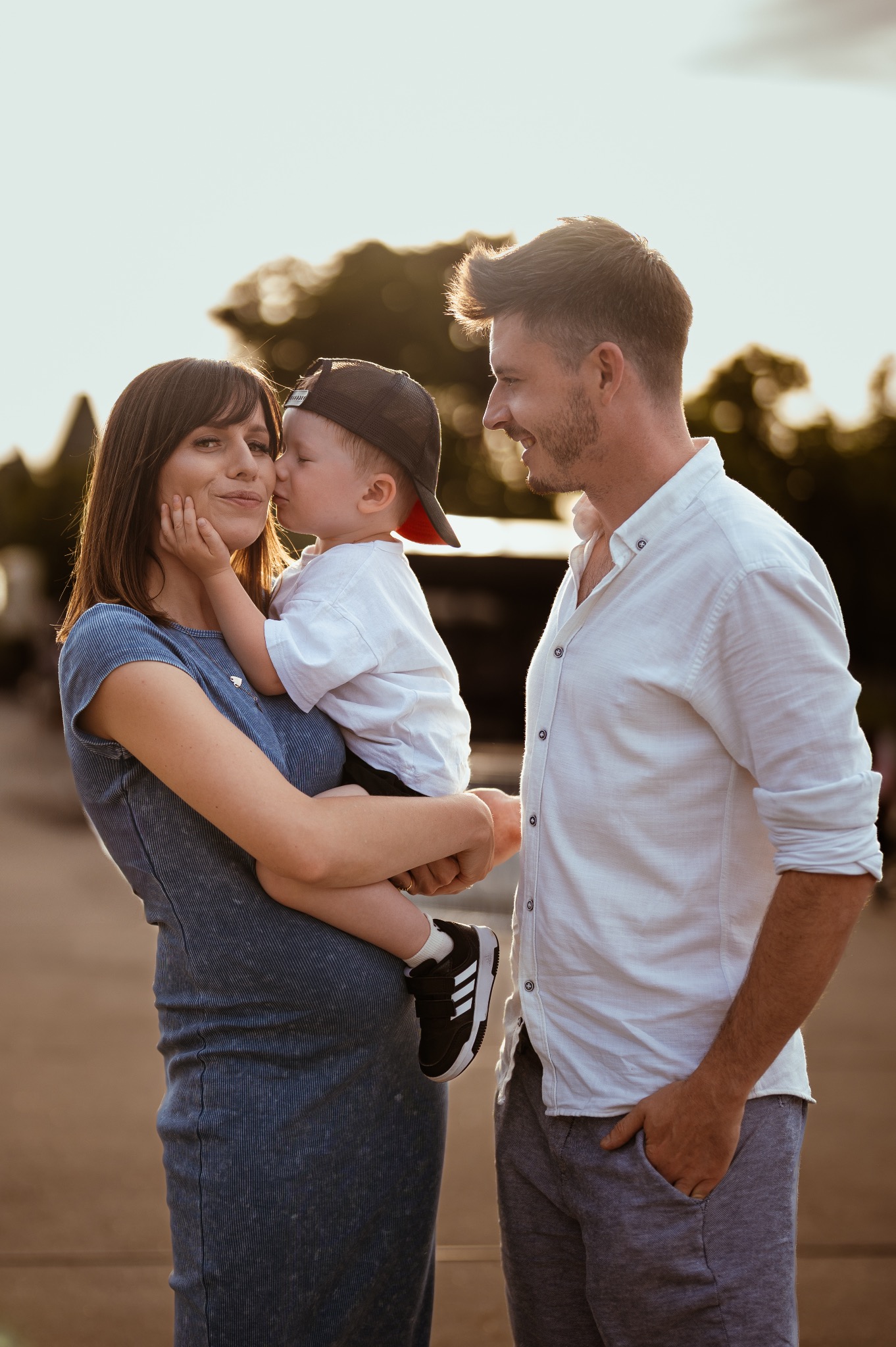 Uśmiechnięta, brzemienna kobieta trzyma synka w czapce, który całuje ją w policzek. Obok stoi mężczyzna w białej koszuli. Ciepłe, naturalne światło.