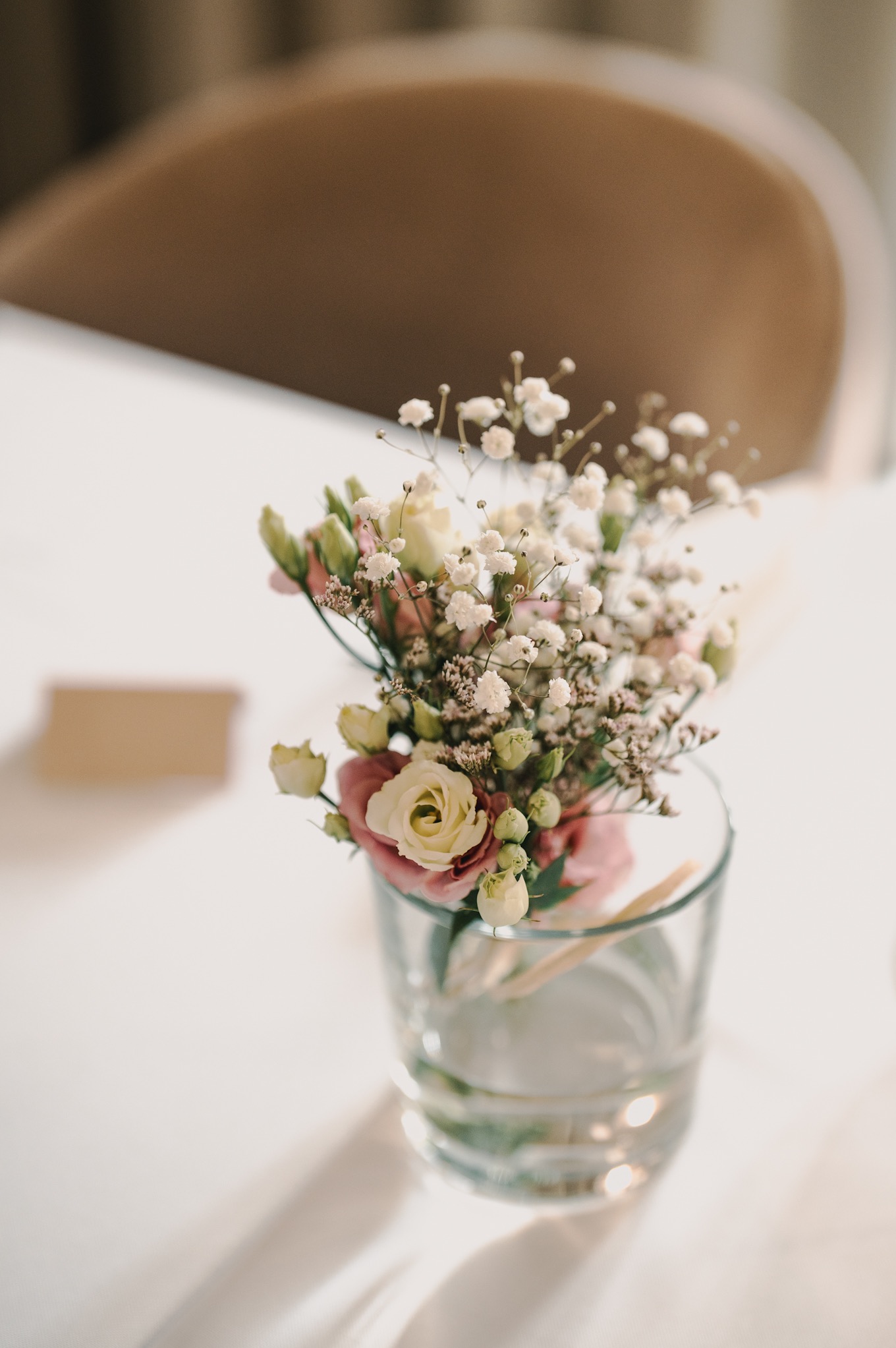 Delikatny bukiecik z białych i różowych kwiatów w szklanym wazonie na stole z kartą w tle, prawdopodobnie dekoracja stołu weselnego w ciepłym oświetleniu.