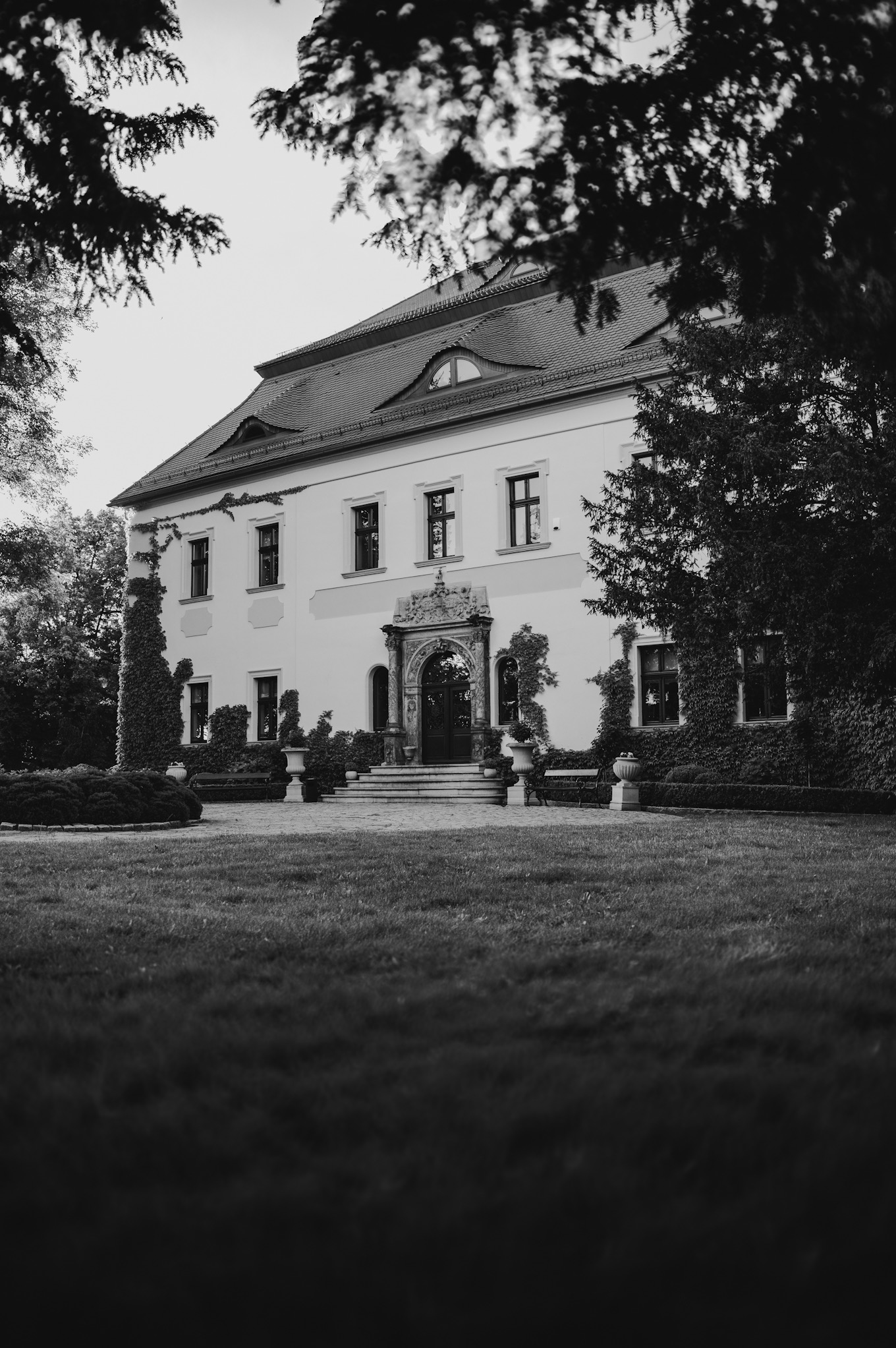 Czarno-białe zdjęcie fasady okazałego, dwupiętrowego budynku z dekoracyjnym portalem wejściowym, porośniętego bluszczem, otoczonego zielenią.