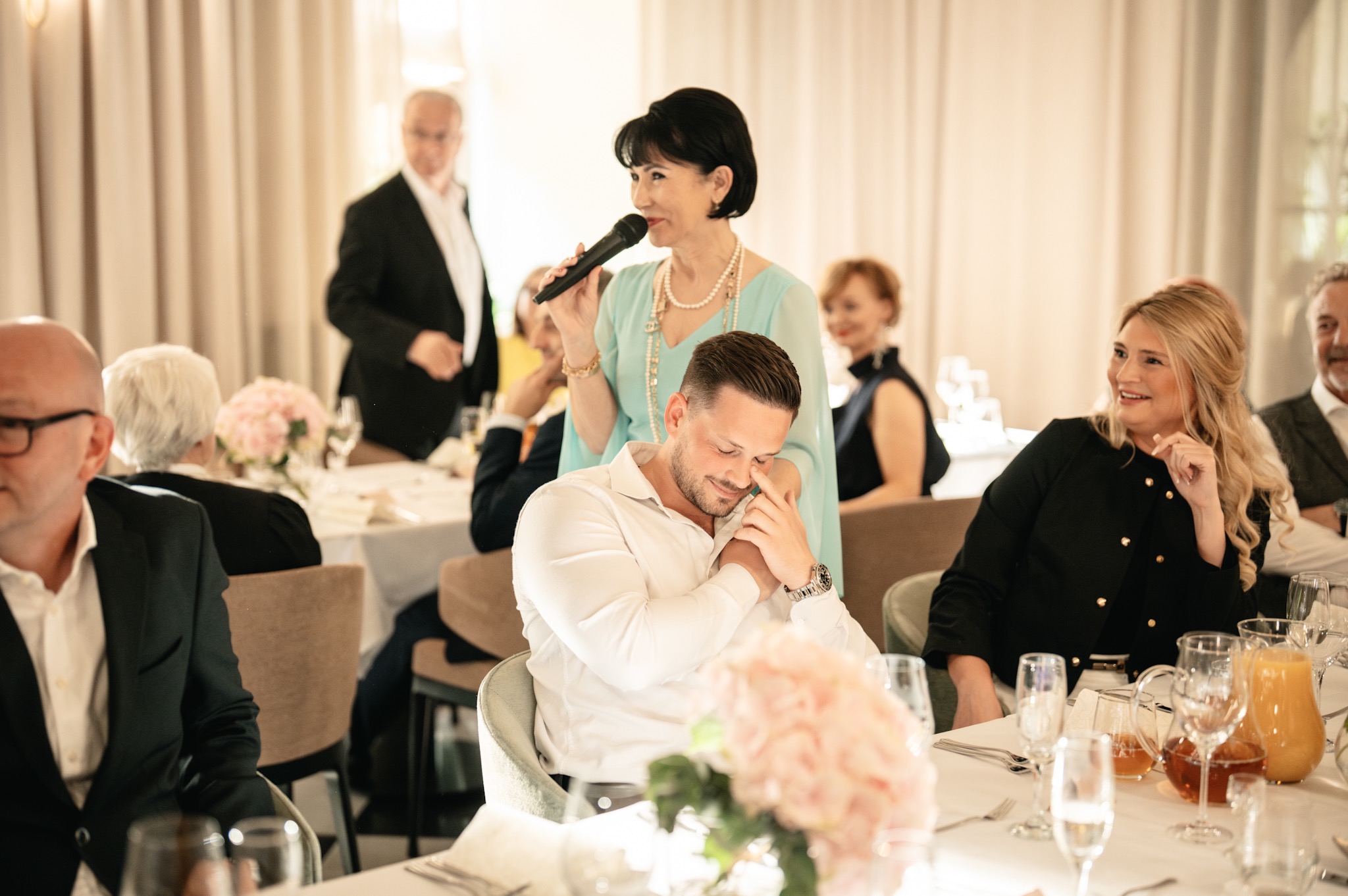Kobieta z mikrofonem przemawia do mężczyzny siedzącego przy stole podczas uroczystości. W tle goście i nakryty stół.
