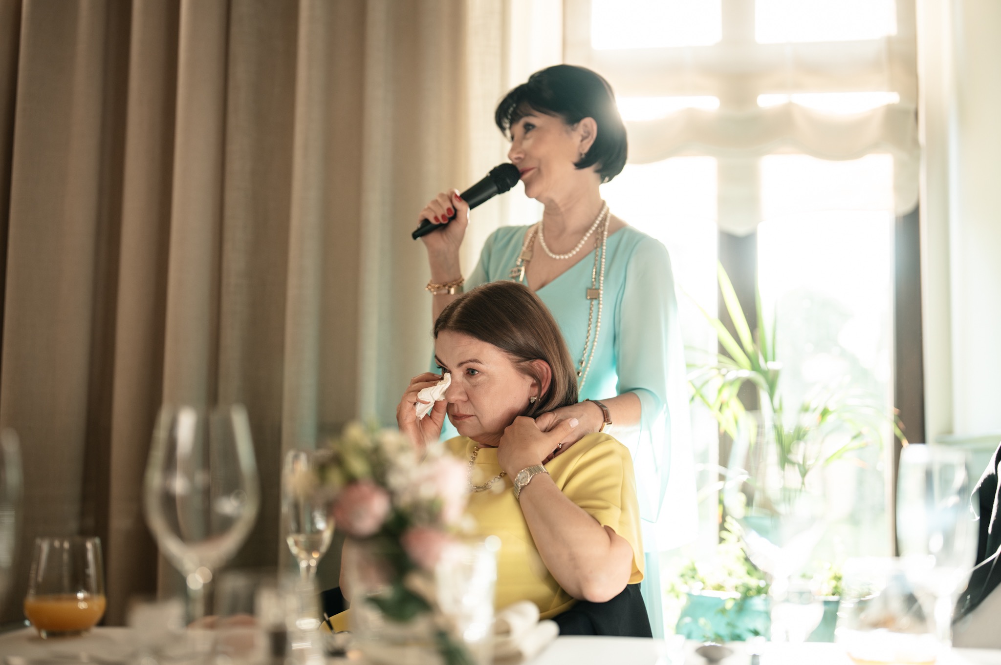 Kobieta z mikrofonem przemawia, a druga kobieta ociera łzy wzruszenia. Ujęcie z bliska, w tle okno i zasłony, widoczne kieliszki i bukiet kwiatów na stole.