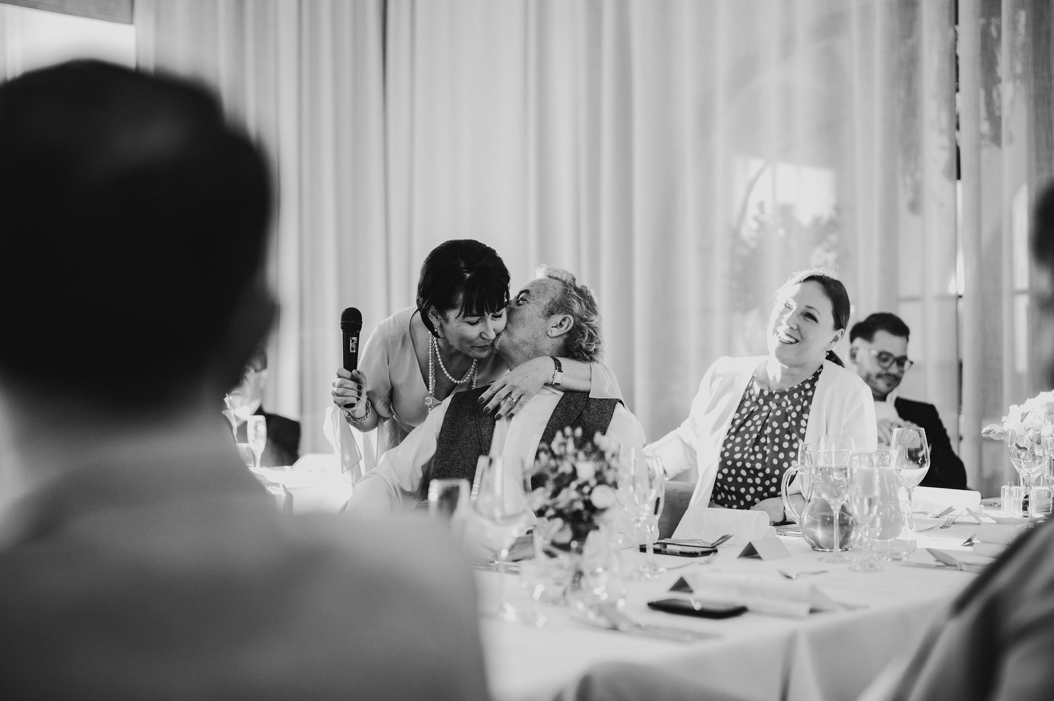 Czarno-białe zdjęcie z wesela: kobieta z mikrofonem całuje pana młodego przy stole, goście weselni obserwują z uśmiechem. Romantyczna atmosfera.