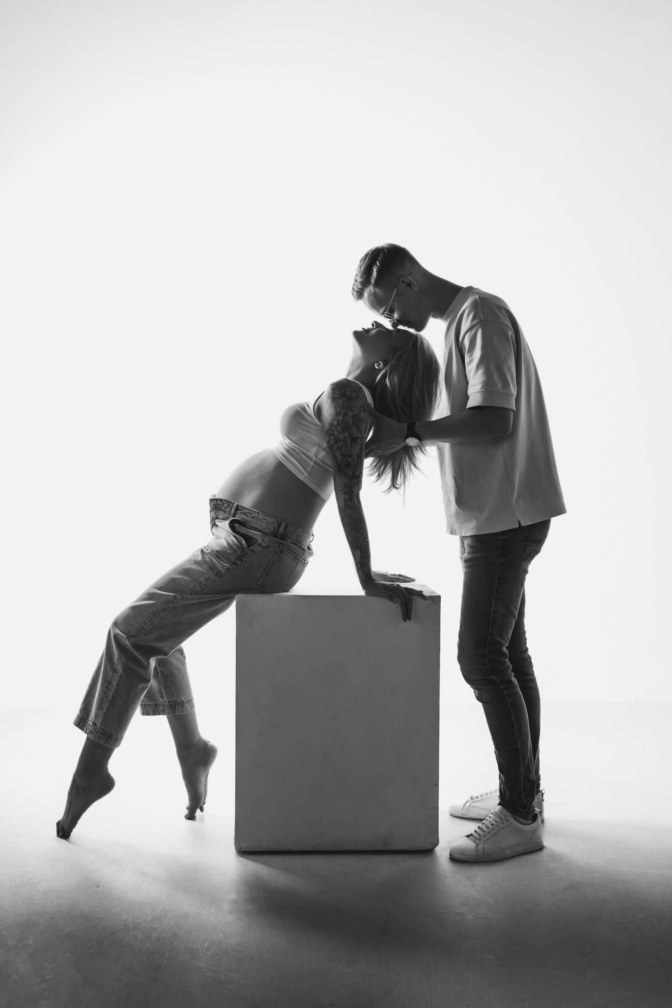 Czarno-białe zdjęcie pary: przyszła mama z tatuażem na ramieniu opiera się o biały sześcian, a mężczyzna w okularach czule dotyka jej włosów. Studio, delikatne światło.