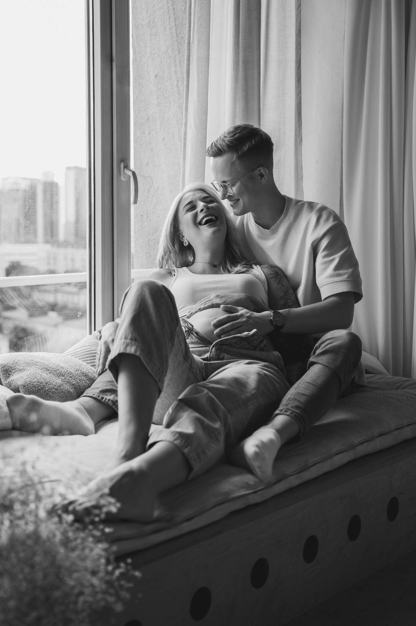 Czarno-białe zdjęcie uśmiechniętej kobiety w ciąży leżącej na parapecie z partnerem, który trzyma ręce na jej brzuchu. W tle okno z widokiem na miasto.