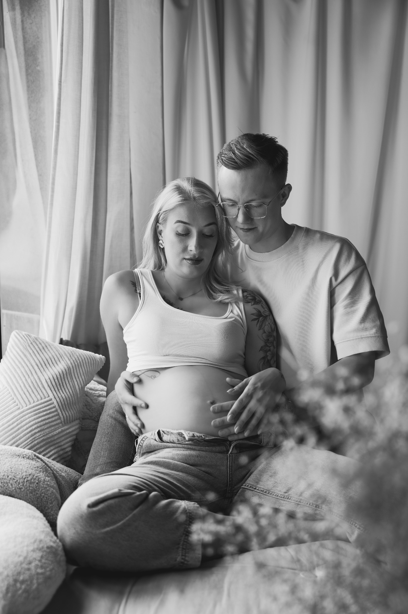 Czarno-białe zdjęcie: przyszła mama z tatą siedzą na kanapie, tuląc brzuszek. Kobieta ma tatuaż na ramieniu i brzuchu. W tle zasłony i kwiaty.