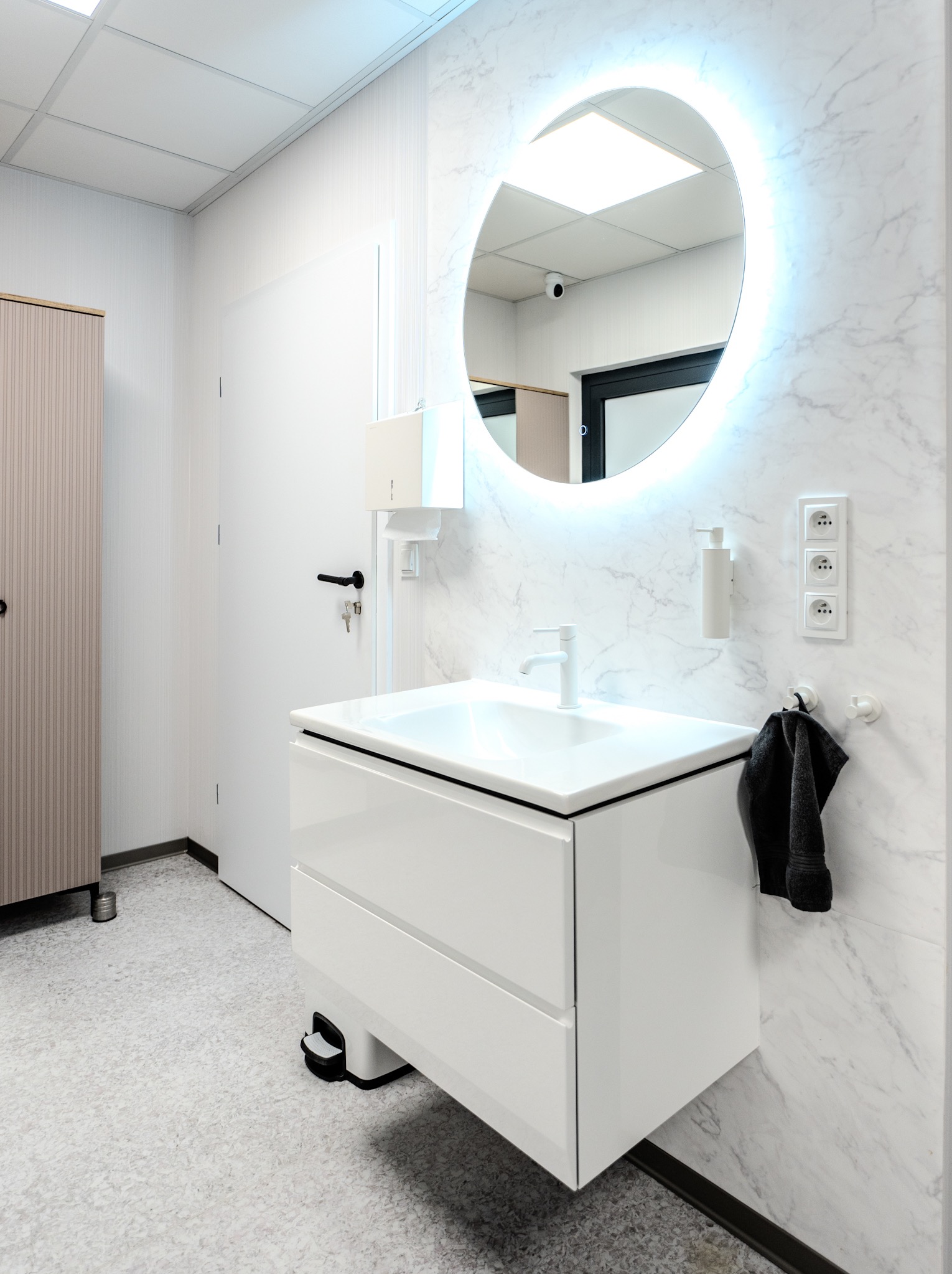 Nowoczesna łazienka w bieli z okrągłym lustrem LED, marmurową ścianą i minimalistyczną umywalką z szafką. Widoczny dozownik mydła i czarny ręcznik.