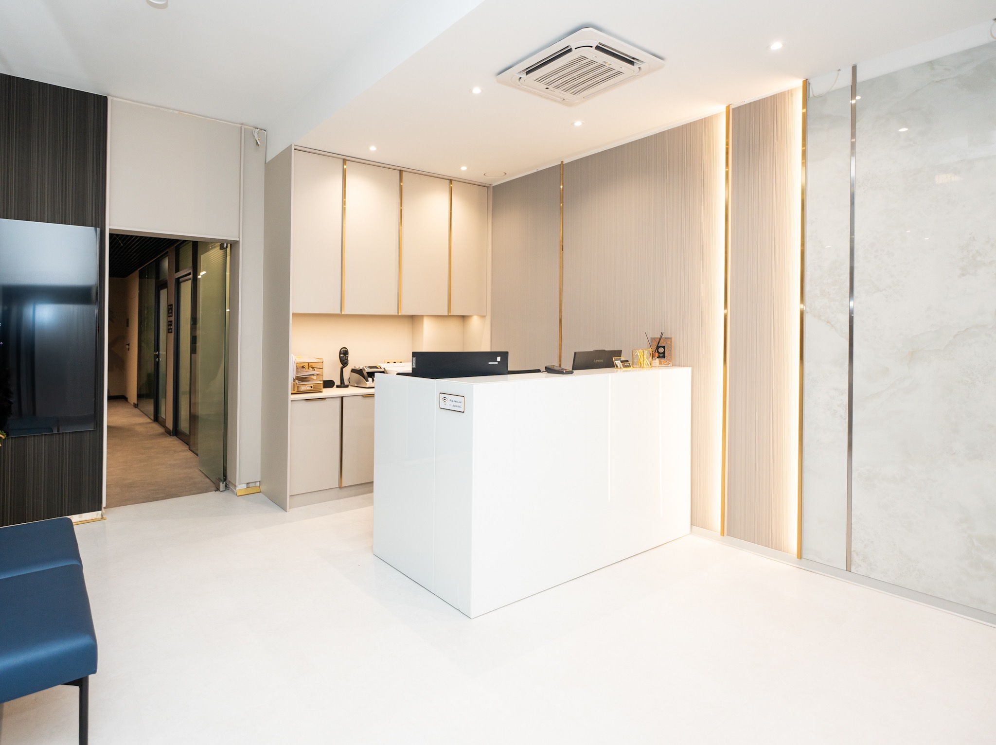 Nowoczesne wnętrze kliniki z białą recepcją, beżowymi szafkami i oświetleniem LED. Minimalistyczny design z elementami dekoracyjnymi i jasną podłogą.