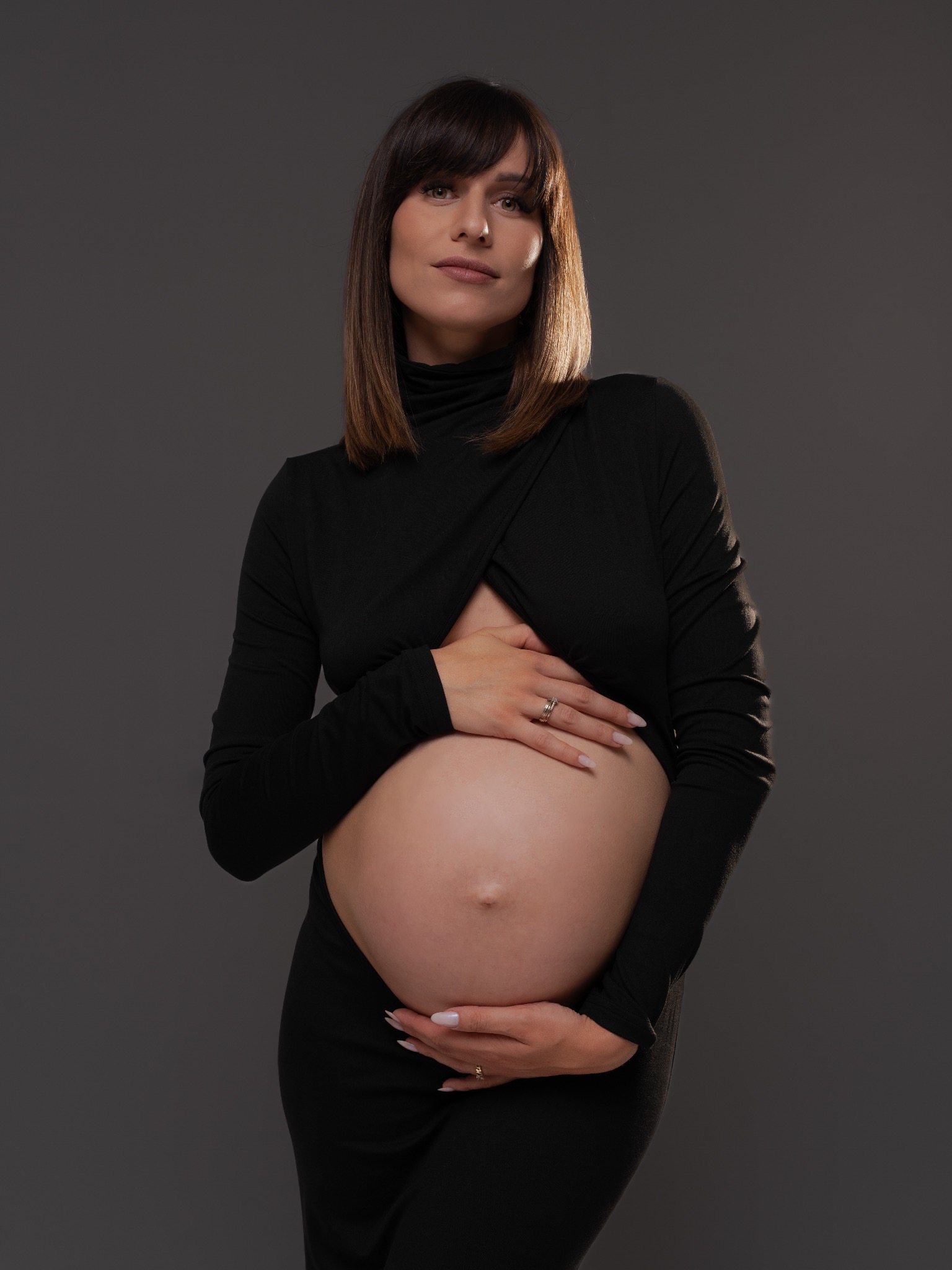 Elegancka sesja ciążowa: Kobieta w czarnej sukni z golfem, z rękami na brzuchu, na stonowanym szarym tle, w studiu, z delikatnym oświetleniem.