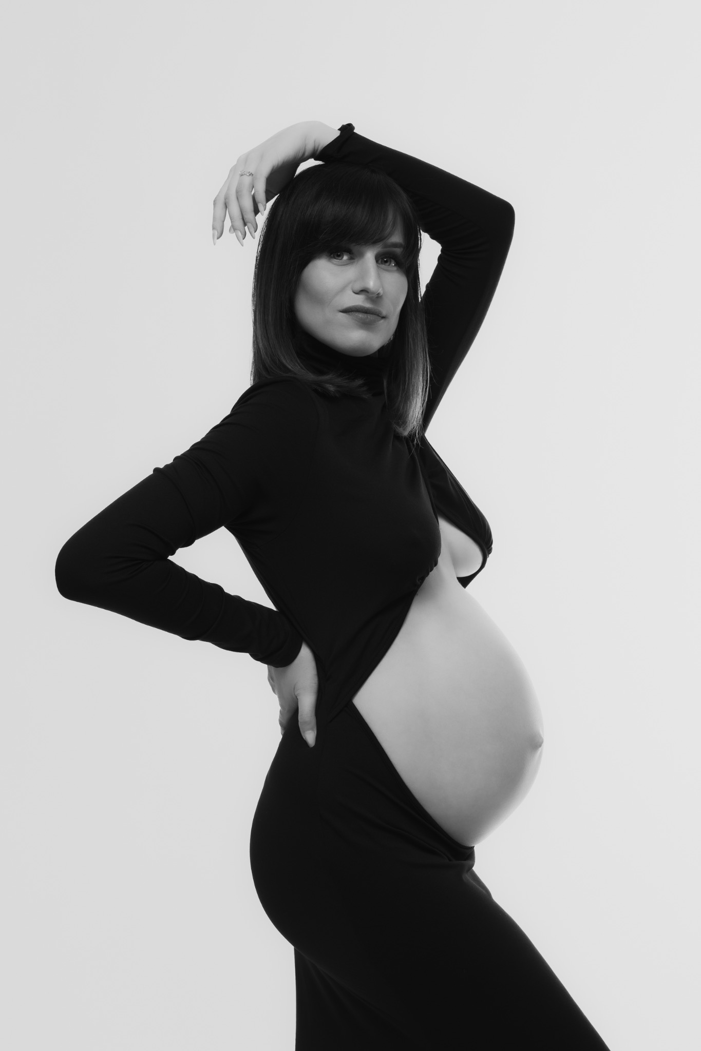 Monochromatyczne zdjęcie kobiety w ciąży, pozującej w czarnym body z golfem, z ręką uniesioną nad głową i dłonią na biodrze, na jasnym tle. Subtelny makijaż, wyraziste spojrzenie.