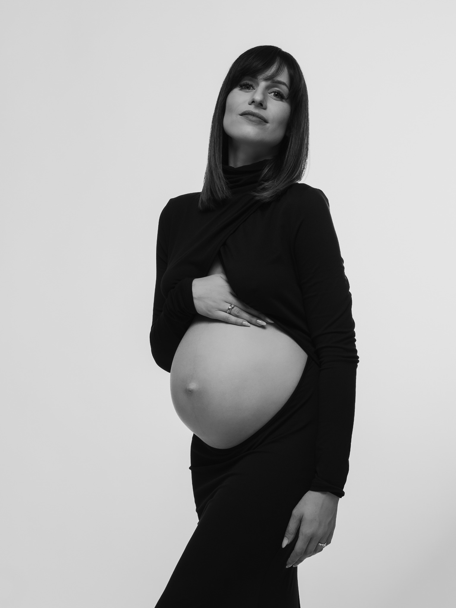 Elegancka, czarno-biała sesja ciążowa: kobieta w czarnej sukience, z ręką na brzuchu, patrzy w górę. Minimalistyczne tło, skupienie na emocjach przyszłej mamy.