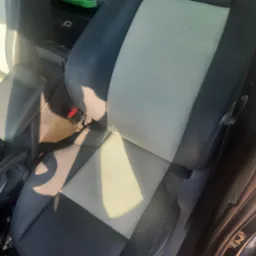 Tapicerowane, dwukolorowe siedzenie samochodowe z perforowaną jasną wstawką i ciemnoszarymi bokami, widoczny fragment czerwonego zapięcia pasa bezpieczeństwa.