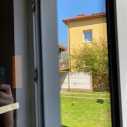 Widok z okna na żółty dom z czerwonym dachem i zielony trawnik z piłką, odbicie ręki z telefonem na szybie.