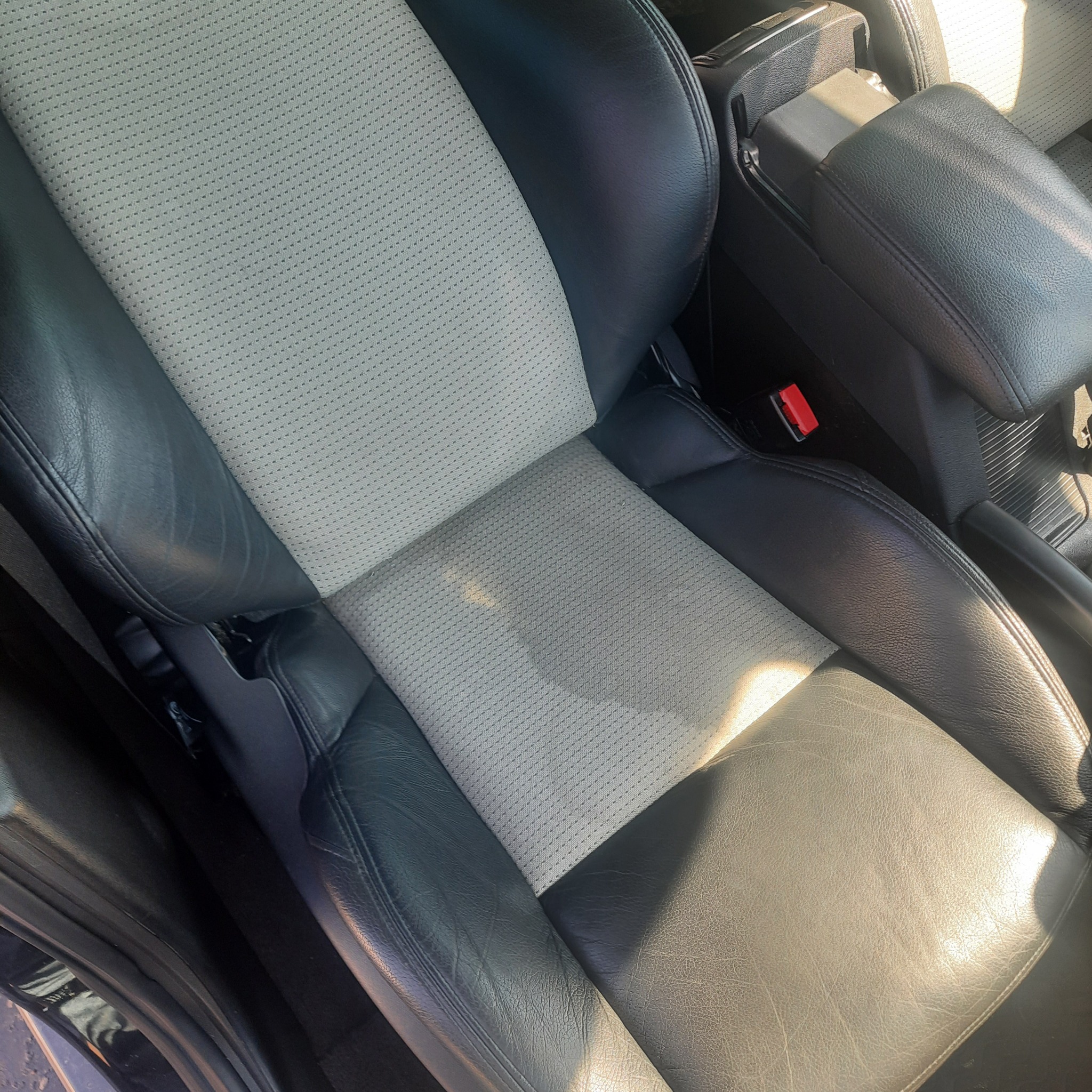 Detail of a two-tone car seat featuring a perforated grey fabric center panel and black leather bolsters, showing wear and sunlight glare.