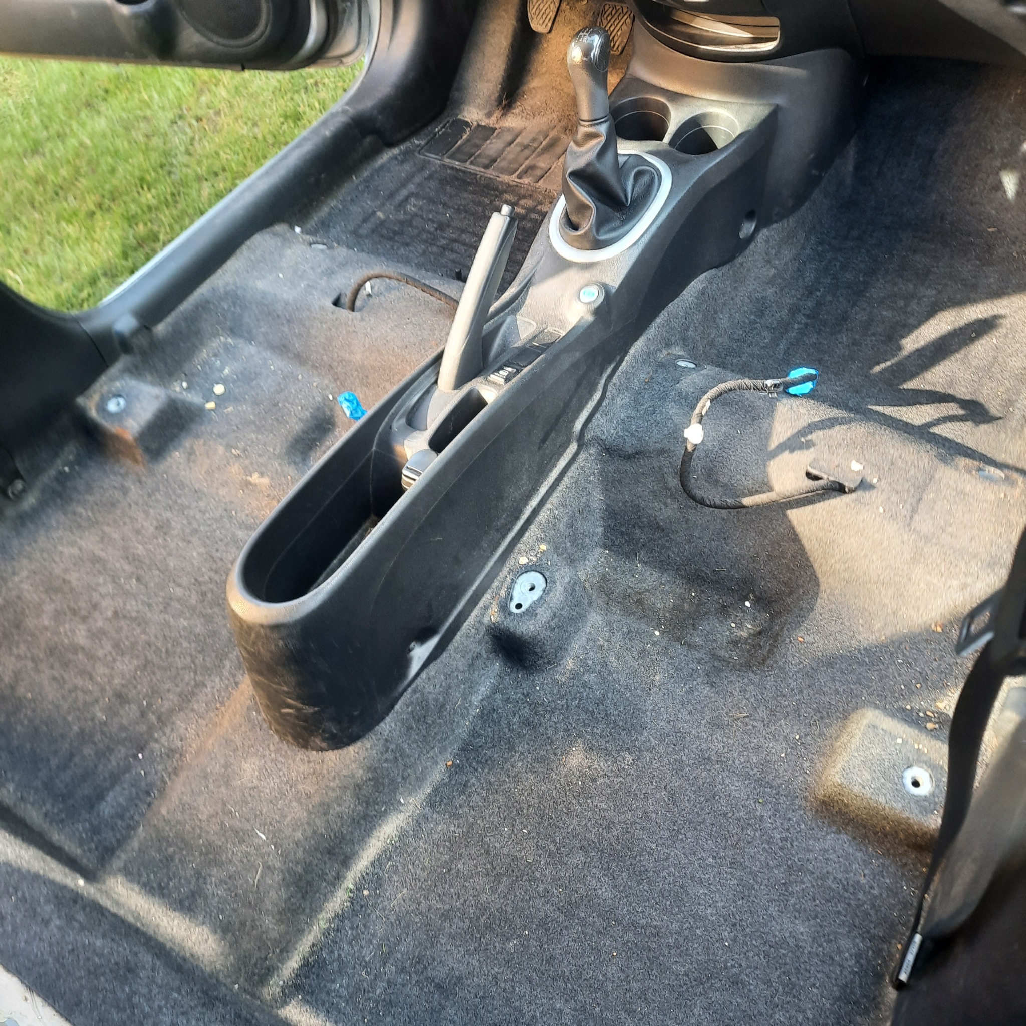 Wnętrze samochodu z wyjętymi fotelami, ukazujące brudną, szarą tapicerkę podłogową przed czyszczeniem, z widocznymi okruchami i zabrudzeniami, oraz odsłoniętymi przewodami.