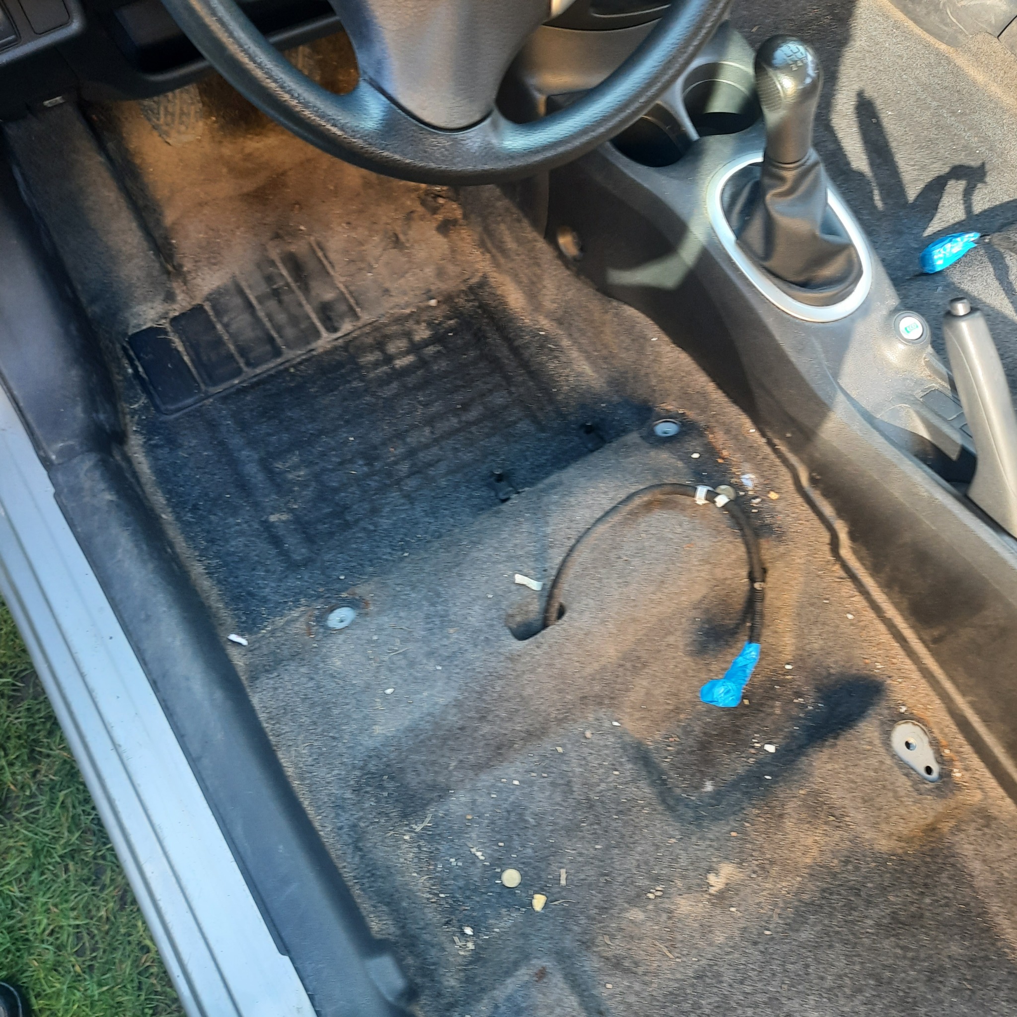 Brudna podłoga samochodu z widocznymi okruchami i zanieczyszczeniami, z czarnym dywanikiem i elementami wnętrza pojazdu, takimi jak kierownica i dźwignia zmiany biegów.