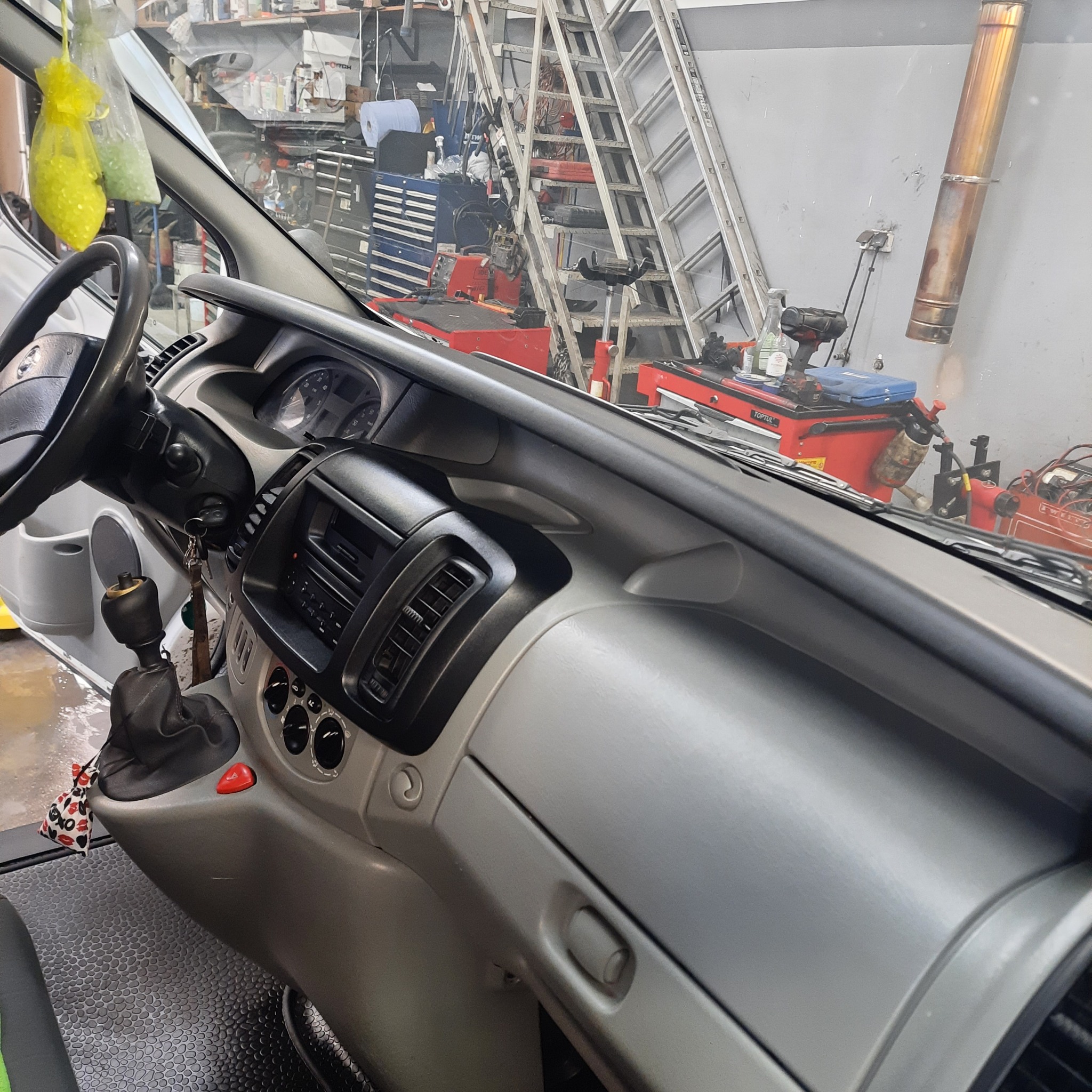 Wnętrze kabiny busa z widoczną deską rozdzielczą, kierownicą i dźwignią zmiany biegów. Na przedniej szybie wiszą zapachowe woreczki z koralikami. W tle widoczne wyposażenie warsztatu samochodowego,...