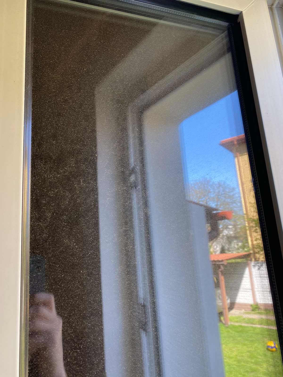 Bardzo brudne okno z widoczną warstwą kurzu i pyłu, przez które widać zarys budynku i niebieskie niebo.