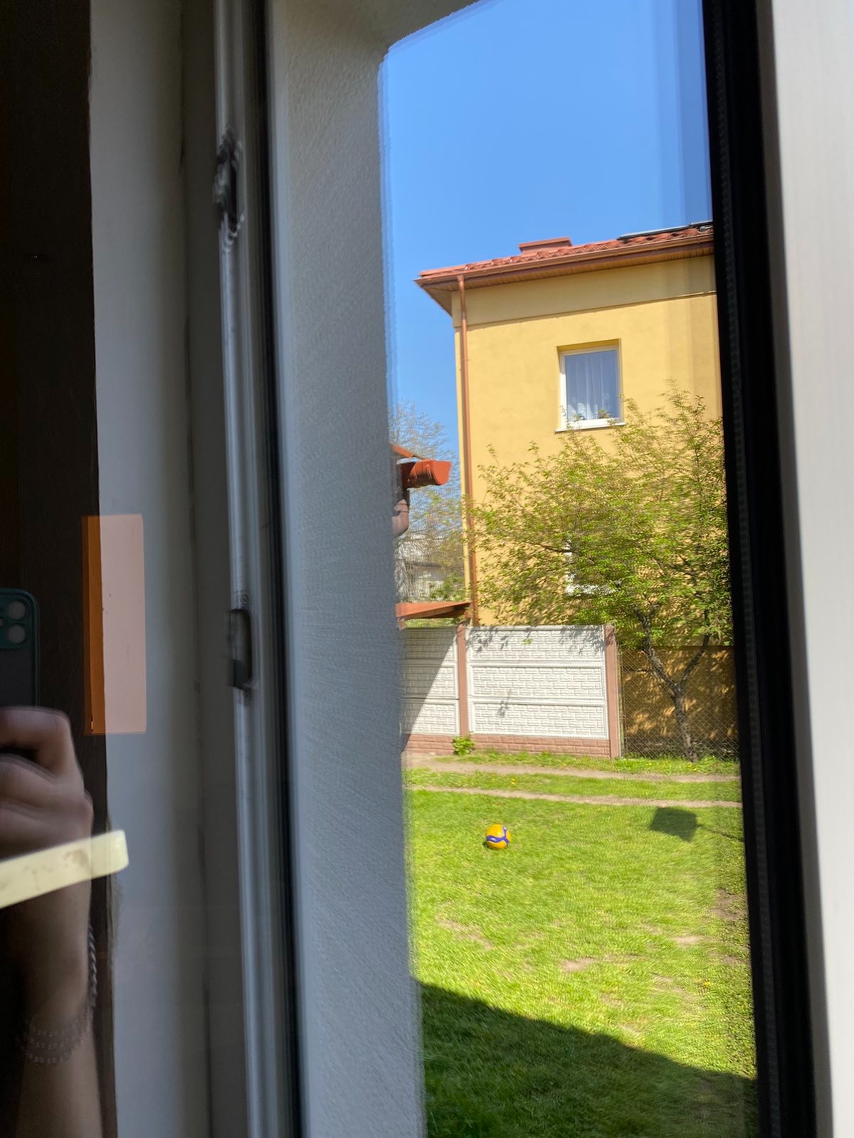 Widok z okna na żółty dom z czerwonym dachem i zielony trawnik z piłką, odbicie ręki z telefonem na szybie.