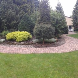 Haus&Garten - Ogród z krętą ścieżką z czerwonej kostki brukowej, otoczony zielonym trawnikiem, ozdobnymi krzewami i wysokimi drzewami iglastymi. Białe, metalowe krzesła i stolik w prawym dolnym rogu.