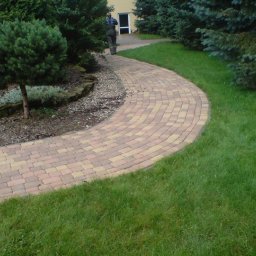 Haus&Garten - Zakrzywiony chodnik z kostki brukowej w odcieniach beżu i brązu, otoczony trawnikiem i rabatą z ozdobnymi krzewami, z sylwetką osoby w tle.