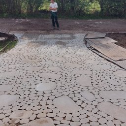 Haus&Garten - Ścieżka z nieregularnych, jasnoszarych kamieni ułożona w ogrodzie, widoczny pracownik w oddali i płyty ochronne na ziemi.