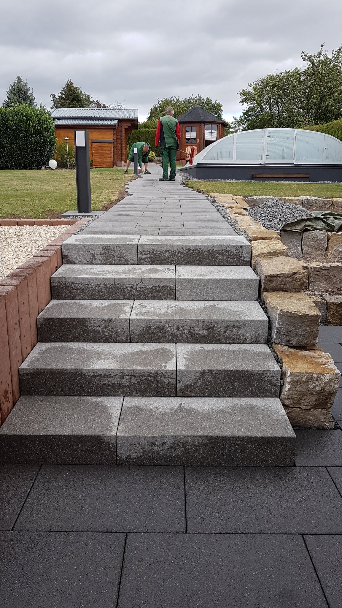 Schody z szarych, prostokątnych bloków kostki brukowej prowadzące przez ogród, dwóch pracowników w zielonych kombinezonach przy pracy, w tle drewniane budynki ogrodowe i zadaszony basen.