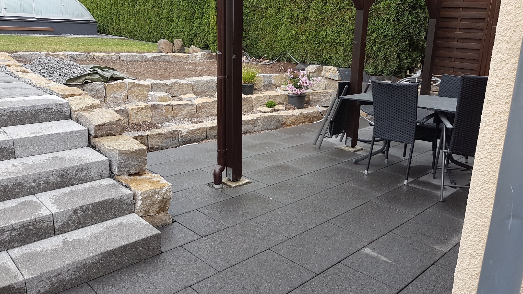 Nowoczesny taras z ciemnoszarej kostki brukowej, schody z jasnych bloków kamiennych, rabata z kamieni i roślin w tle, zestaw mebli ogrodowych pod zadaszeniem.