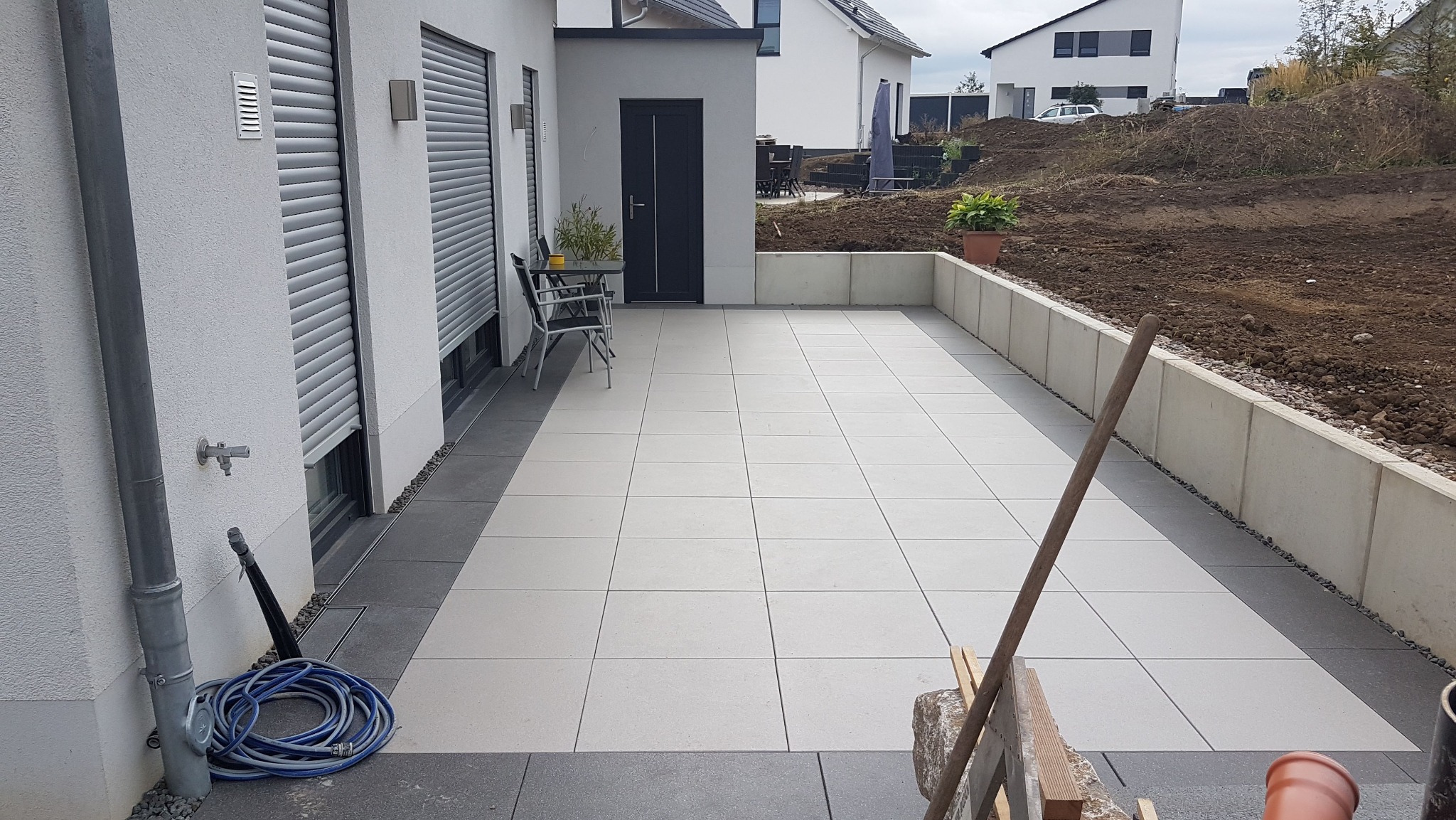 Nowoczesny taras wyłożony jasnymi, kwadratowymi płytami, z ciemnoszarym obramowaniem, widok na przygotowany teren pod ogród i część domu z roletami.