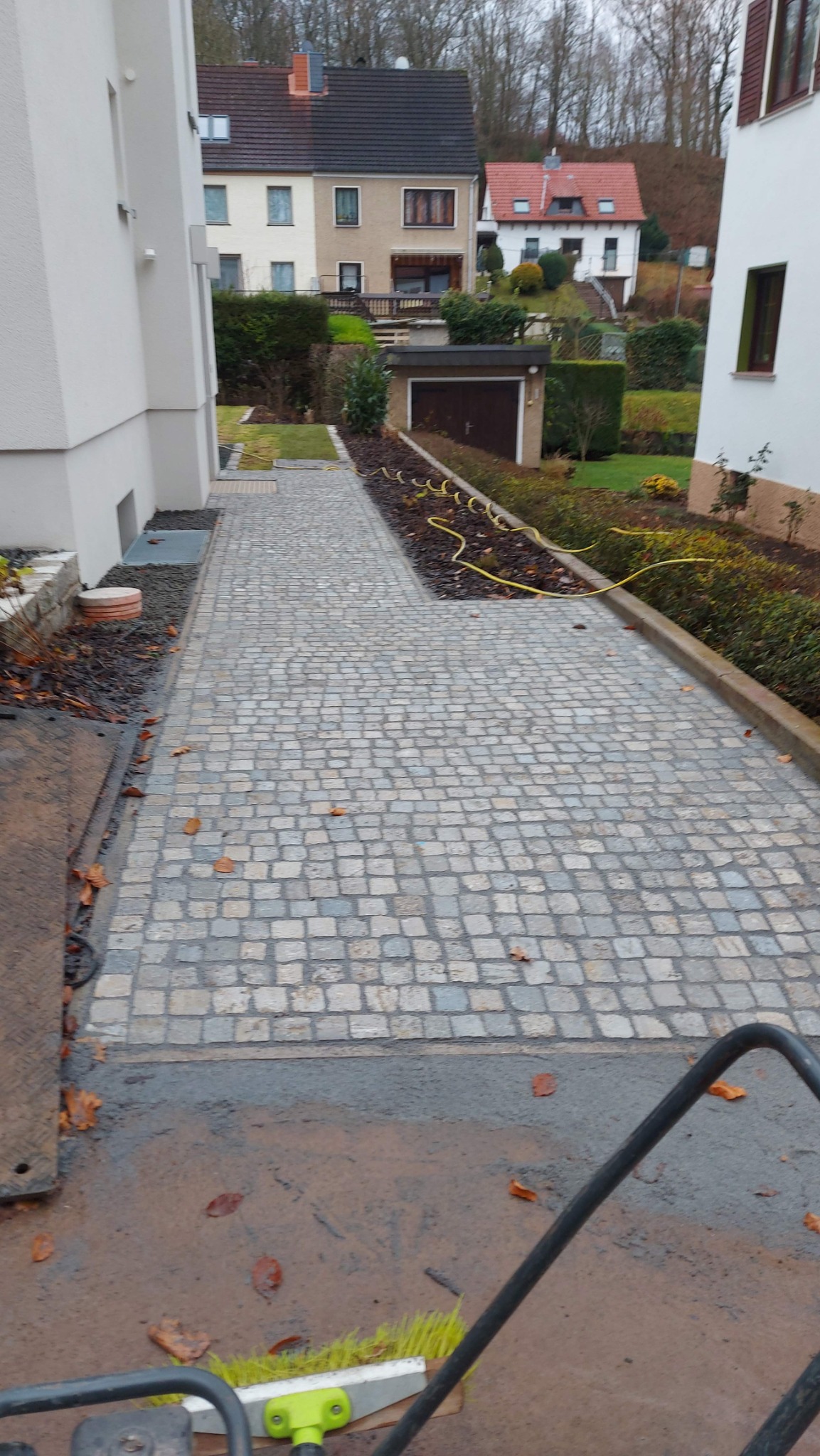Ścieżka i podjazd z kostki granitowej ułożone między dwoma budynkami mieszkalnymi, widoczny wózek z miotłą do zamiatania.