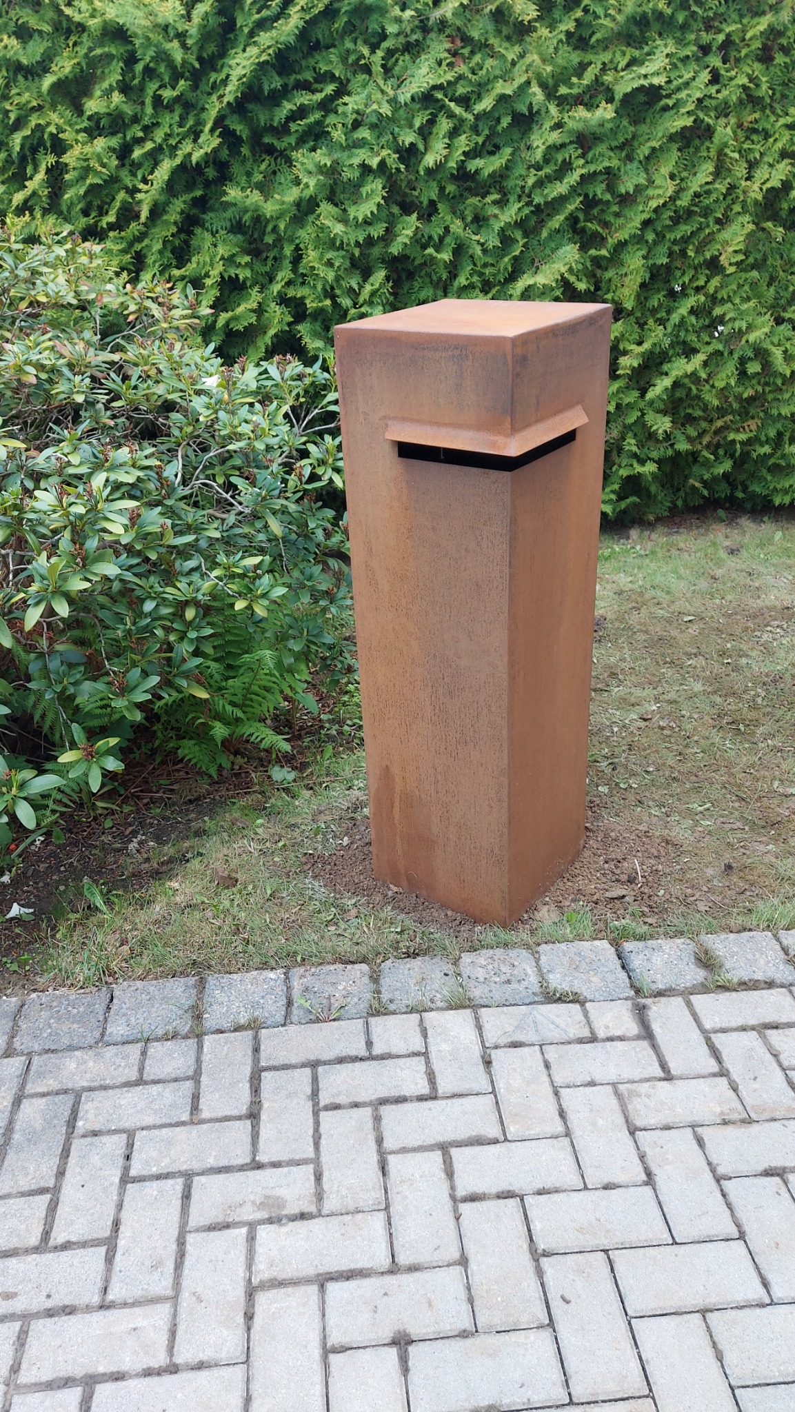 Rdzawa, metalowa skrzynka na listy o kubistycznym kształcie, umieszczona na trawniku obok ścieżki z kostki brukowej, z tłem w postaci zielonych krzewów.