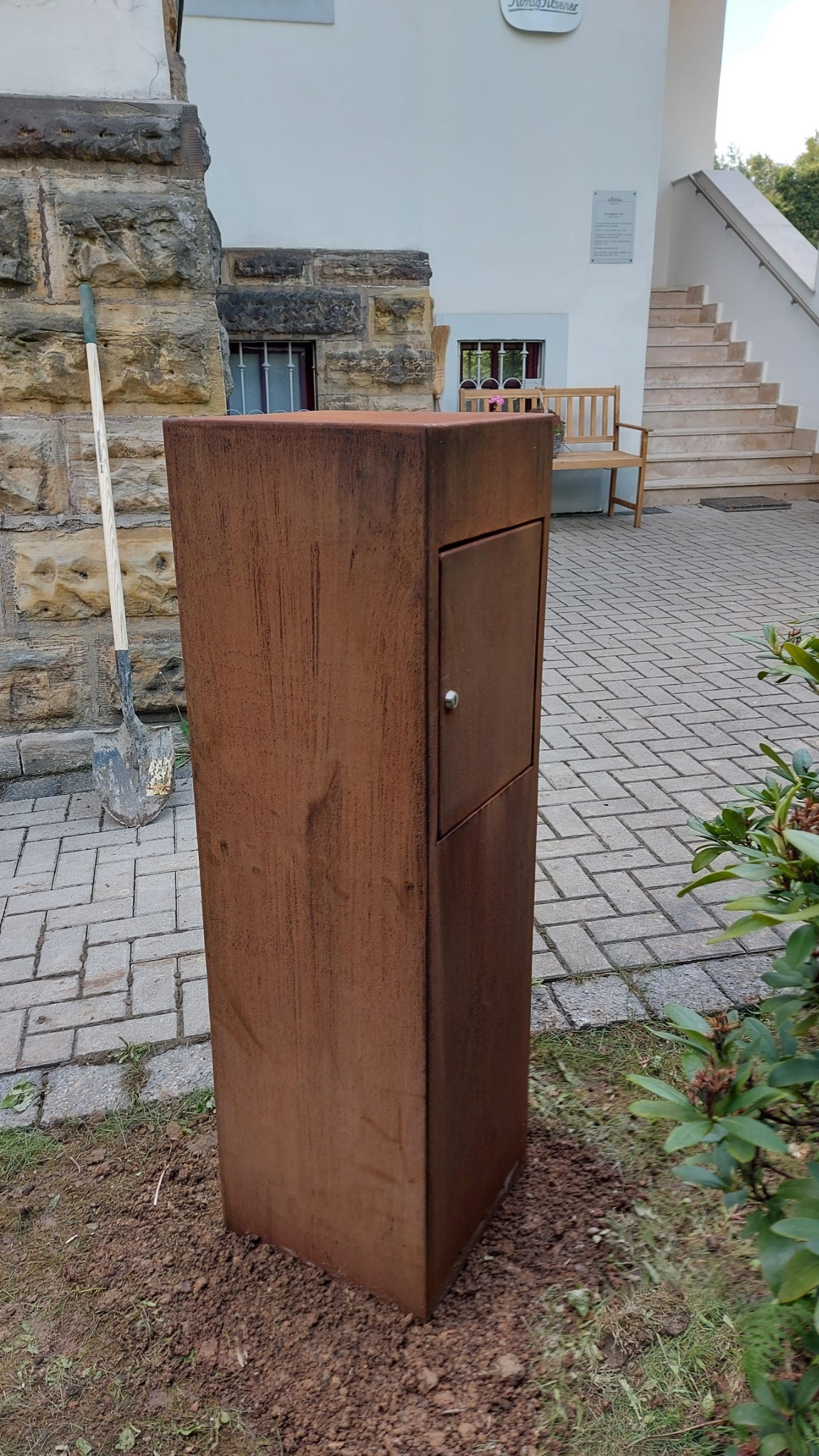 Rdzewna, metalowa skrzynka z drzwiczkami i zamkiem, posadowiona w ziemi obok chodnika z kostki, łopata oparta o kamienną ścianę w tle.
