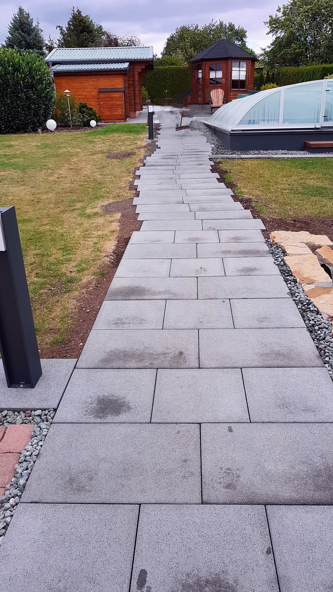 Prosta ścieżka z szarych, prostokątnych płyt prowadząca przez trawnik w kierunku drewnianych budynków ogrodowych i zadaszonego basenu, z widocznymi śladami stóp na niektórych płytach.